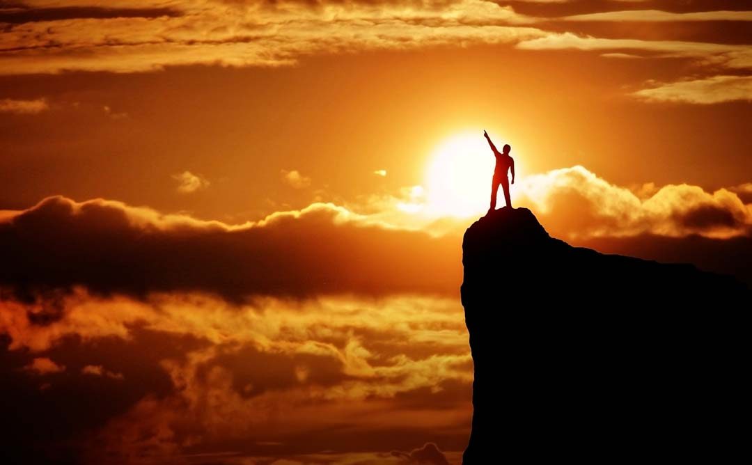 Silhouetted figure standing at the top of a mountain pointing to the sky